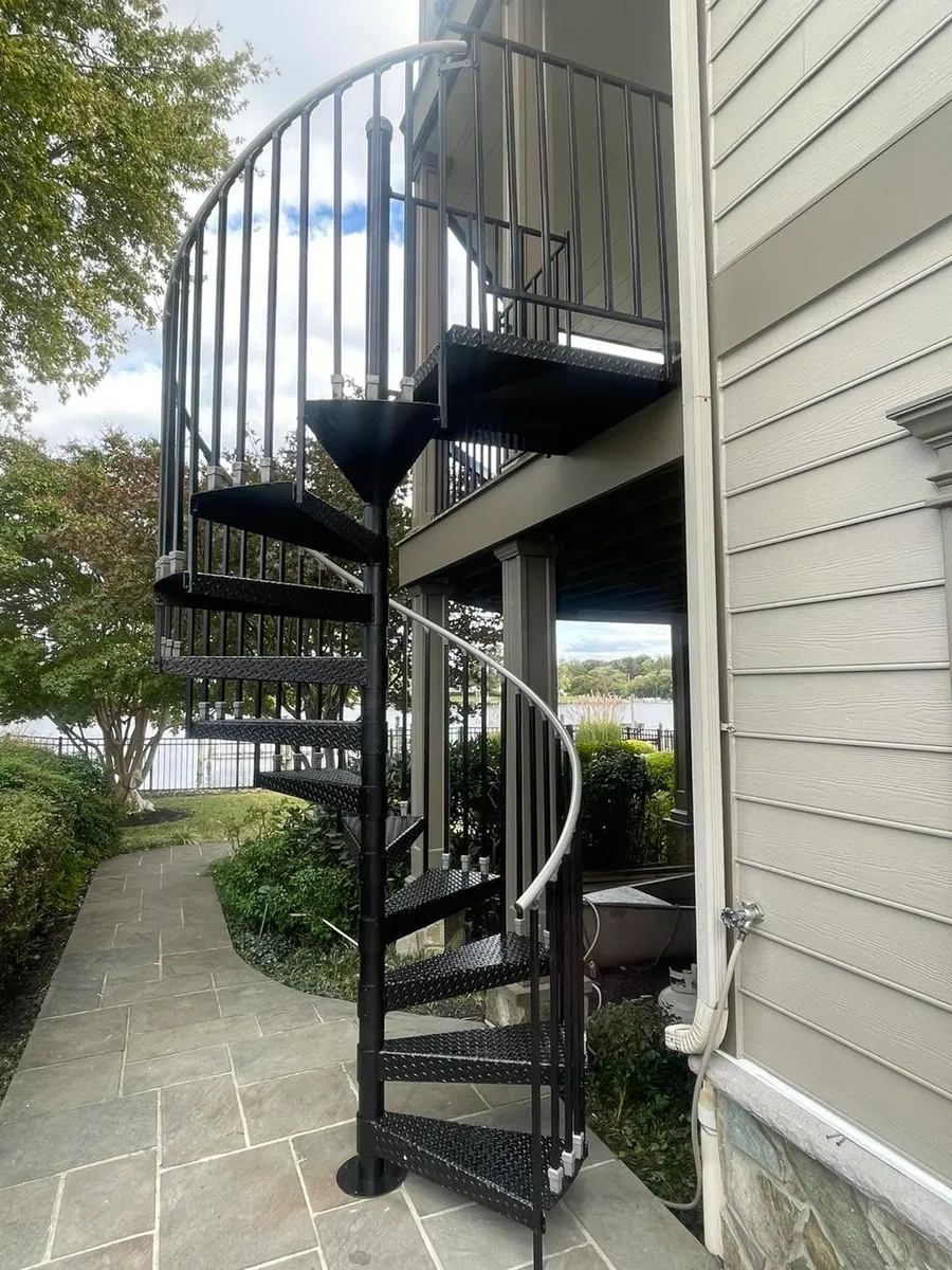 Freshly painted black metal spiral staircase on a flagstone patio with water and gardens behind.